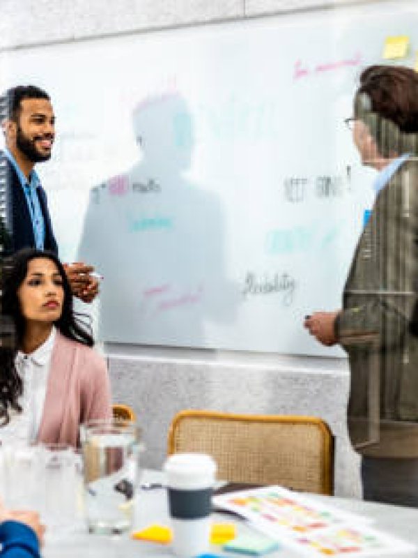 Diverse male coaches teaching employees kanban project planning technique in conference room.  Multiracial business people putting sticky notes on whiteboard